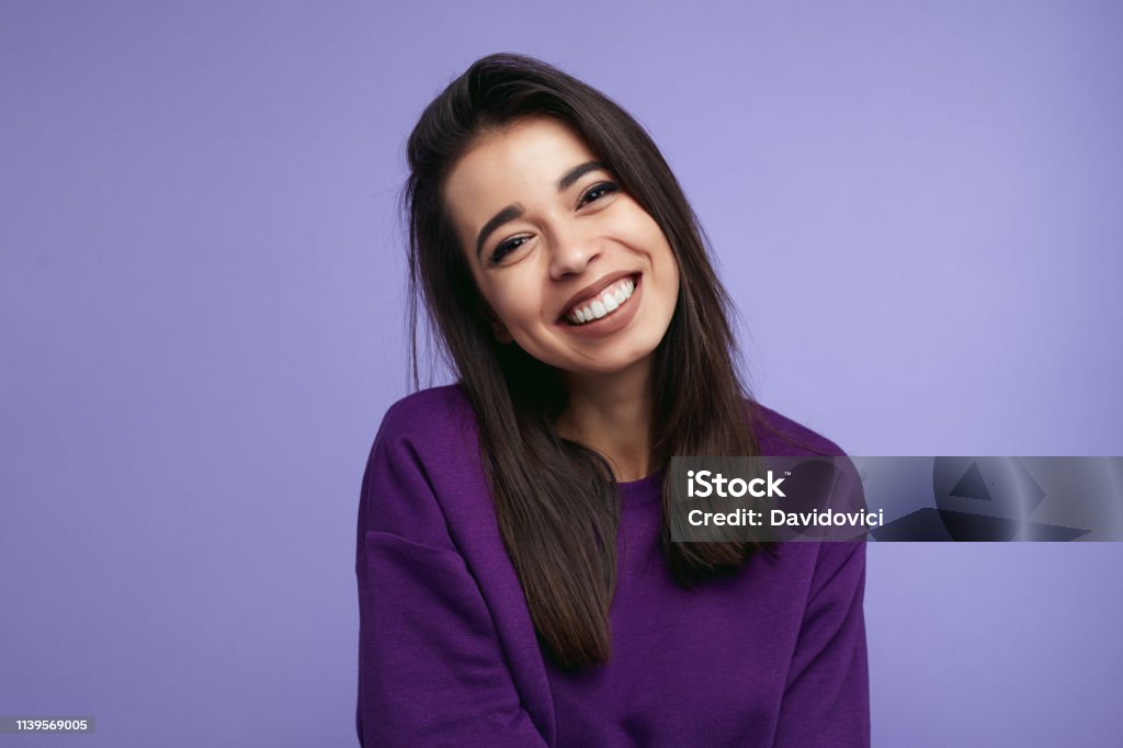 Portrait of pretty adult girl, broad smile, wears purple sweatshirt, tilts head and looks with joy, dressed casually, isolated over violet background